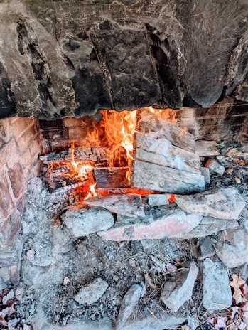 A rustic stone fireplace with a vibrant fire burning, surrounded by ash and charred logs. The interior is lined with bricks and the stone exterior appears weathered and rugged.