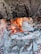 A rustic stone fireplace with a vibrant fire burning, surrounded by ash and charred logs. The interior is lined with bricks and the stone exterior appears weathered and rugged.