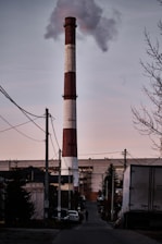A large industrial smokestack emits smoke under a cloudy sky in a suburban or industrial area. The scene includes several buildings, utility poles, and cables. A few vehicles and a person are visible in the foreground along the street.