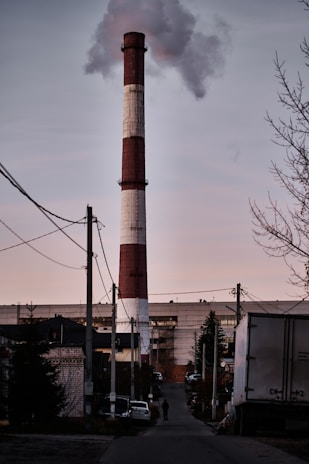 A large industrial smokestack emits smoke under a cloudy sky in a suburban or industrial area. The scene includes several buildings, utility poles, and cables. A few vehicles and a person are visible in the foreground along the street.