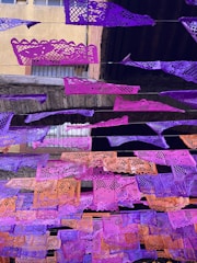 Colorful paper banners with intricate cut-out designs are strung across an outdoor space. The banners, primarily in shades of purple and orange, contrast with the aged stone and brick structure behind them. Windows with bars can be seen in the background, indicating an urban setting.
