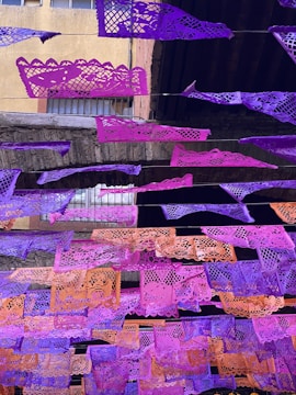 Colorful paper banners with intricate cut-out designs are strung across an outdoor space. The banners, primarily in shades of purple and orange, contrast with the aged stone and brick structure behind them. Windows with bars can be seen in the background, indicating an urban setting.