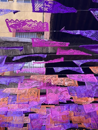 Colorful paper banners with intricate cut-out designs are strung across an outdoor space. The banners, primarily in shades of purple and orange, contrast with the aged stone and brick structure behind them. Windows with bars can be seen in the background, indicating an urban setting.