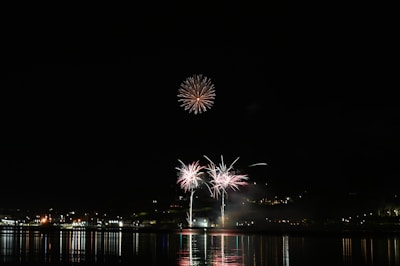 Fireworks illuminate the night sky above a waterfront, reflecting vibrant colors onto the water below. The display includes bursts of light and color in various patterns, set against a backdrop of distant city lights.