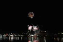 Fireworks illuminate the night sky above a waterfront, reflecting vibrant colors onto the water below. The display includes bursts of light and color in various patterns, set against a backdrop of distant city lights.