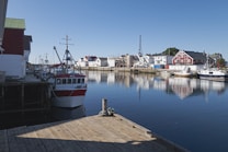 A tranquil harbor scene features several fishing boats docked along a wooden pier. Traditional-style buildings with red and white facades line the water, creating a picturesque reflection on the calm water surface. The clear blue sky indicates a sunny day, contributing to the serene atmosphere.