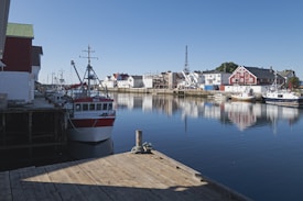 A tranquil harbor scene features several fishing boats docked along a wooden pier. Traditional-style buildings with red and white facades line the water, creating a picturesque reflection on the calm water surface. The clear blue sky indicates a sunny day, contributing to the serene atmosphere.