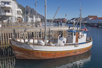 A wooden fishing boat is docked at a pier in a picturesque coastal town. The boat has a white upper section with a natural wood finish on the lower part. A vibrant blue roof caps the cabin, and life buoys are visible on the sides. The background features charming white and red buildings under a clear blue sky, with rugged mountains in the distance.