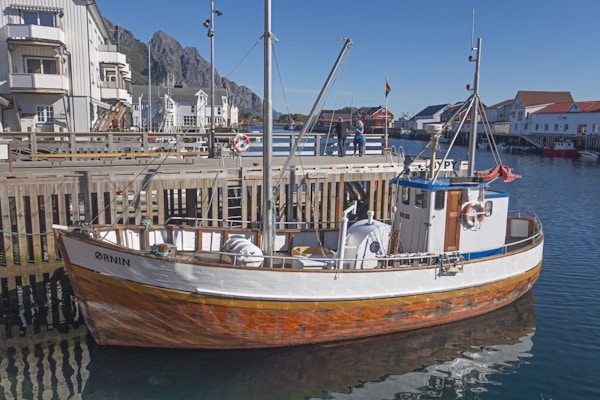 A wooden fishing boat is docked at a pier in a picturesque coastal town. The boat has a white upper section with a natural wood finish on the lower part. A vibrant blue roof caps the cabin, and life buoys are visible on the sides. The background features charming white and red buildings under a clear blue sky, with rugged mountains in the distance.
