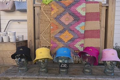 A collection of colorful hats is displayed on glass mannequin heads on a wooden shelf. Behind the hats, there are woven rugs with geometric patterns in vibrant colors, hanging as a backdrop. To the side, several ceramic mugs are arranged on a wooden crate next to what appears to be woven bags hanging from a stand.