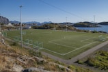 A scenic outdoor football field is positioned against a backdrop of rugged mountains and calm sea. The field is surrounded by a few scattered houses and rocky terrain, with goal posts at each end. The sky is clear and blue, suggesting a nice weather day.