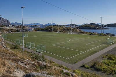 A scenic outdoor football field is positioned against a backdrop of rugged mountains and calm sea. The field is surrounded by a few scattered houses and rocky terrain, with goal posts at each end. The sky is clear and blue, suggesting a nice weather day.