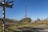 A tall metal communications tower stands on a rocky outcrop in the center of a grassy field. Surrounding the tower are remnants of old wooden structures, some of which appear collapsed. The sky is clear and blue, indicating a sunny day.