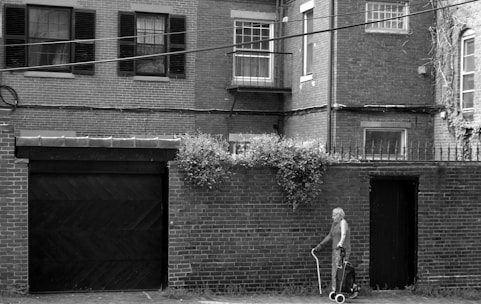 An elderly person walks along a brick-lined street with a wheeled walker. The brick building in the background has multiple windows, some with shutters and a small balcony. Vegetation grows on the wall above a door, adding a touch of greenery.