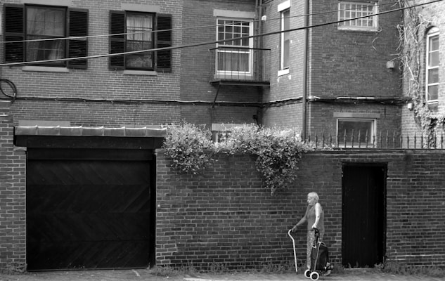 An elderly person walks along a brick-lined street with a wheeled walker. The brick building in the background has multiple windows, some with shutters and a small balcony. Vegetation grows on the wall above a door, adding a touch of greenery.