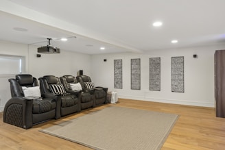 A modern home theater room featuring a row of black, cushioned recliner chairs with pillows. A ceiling-mounted projector is positioned above. The wooden floor is partially covered by a textured rug, and there are decorative panels on the white walls.