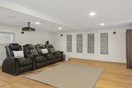 A modern home theater room featuring a row of black, cushioned recliner chairs with pillows. A ceiling-mounted projector is positioned above. The wooden floor is partially covered by a textured rug, and there are decorative panels on the white walls.