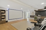 A modern home theater room featuring a large wall-mounted screen and a set of black leather recliner seats with cup holders. To the left, there is a bookshelf with decorative items such as baskets and books. On the right side, a kitchenette with black subway tiles and a mini-fridge is visible. The room has wooden flooring and a neutral-toned rug on the floor. The lighting is recessed, providing a cozy and inviting atmosphere.