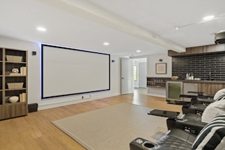 A modern home theater room featuring a large wall-mounted screen and a set of black leather recliner seats with cup holders. To the left, there is a bookshelf with decorative items such as baskets and books. On the right side, a kitchenette with black subway tiles and a mini-fridge is visible. The room has wooden flooring and a neutral-toned rug on the floor. The lighting is recessed, providing a cozy and inviting atmosphere.