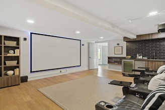 A modern home theater room featuring a large wall-mounted screen and a set of black leather recliner seats with cup holders. To the left, there is a bookshelf with decorative items such as baskets and books. On the right side, a kitchenette with black subway tiles and a mini-fridge is visible. The room has wooden flooring and a neutral-toned rug on the floor. The lighting is recessed, providing a cozy and inviting atmosphere.