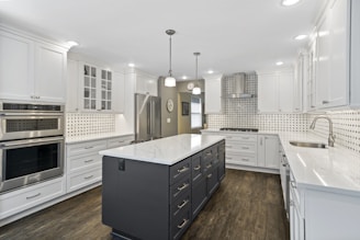 A modern kitchen with a large central island featuring a dark finish and a white marble countertop. Surrounding it are white cabinets and drawers with stainless steel handles. The wall features a stylish black and white tiled backsplash paired with stainless steel appliances, including an oven and refrigerator. Pendant lights hang from the ceiling, illuminating the space, which also has a wooden floor.