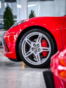 A close-up view of a sleek, modern car wheel with a shiny silver alloy rim and a visible red brake caliper. The car's vivid red body is visible, reflecting light and giving a polished appearance. Background elements include blurred green plants and a bright indoor setting, suggesting a showroom or garage.
