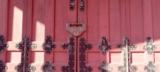 Wooden door painted in a deep pinkish-red shade, adorned with intricate black metal hinges and a central padlock. The door features a symmetrical arrangement of panels, enhanced by the texture of the wood grain and the decorative metalwork.