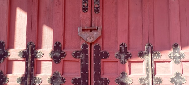 Wooden door painted in a deep pinkish-red shade, adorned with intricate black metal hinges and a central padlock. The door features a symmetrical arrangement of panels, enhanced by the texture of the wood grain and the decorative metalwork.