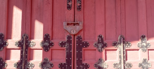 Wooden door painted in a deep pinkish-red shade, adorned with intricate black metal hinges and a central padlock. The door features a symmetrical arrangement of panels, enhanced by the texture of the wood grain and the decorative metalwork.