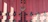 Wooden door painted in a deep pinkish-red shade, adorned with intricate black metal hinges and a central padlock. The door features a symmetrical arrangement of panels, enhanced by the texture of the wood grain and the decorative metalwork.