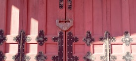 Wooden door painted in a deep pinkish-red shade, adorned with intricate black metal hinges and a central padlock. The door features a symmetrical arrangement of panels, enhanced by the texture of the wood grain and the decorative metalwork.