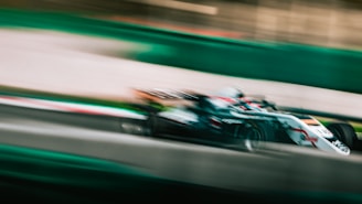 A racing car speeds along a track, with a blur effect emphasizing its high velocity. The car is predominantly white with some red and black accents. The surroundings show motion blur, adding to the sense of speed.