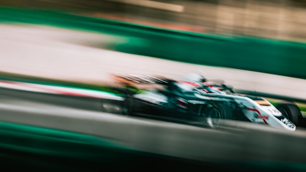A racing car speeds along a track, with a blur effect emphasizing its high velocity. The car is predominantly white with some red and black accents. The surroundings show motion blur, adding to the sense of speed.