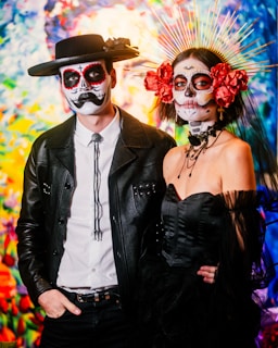 A couple dressed in elaborate Dia de los Muertos costumes with detailed face paint. The man wears a black hat, bolo tie, and a leather jacket, while the woman has a headdress with golden spikes and red flowers. The background is vibrant and colorful, resembling a painted mural.