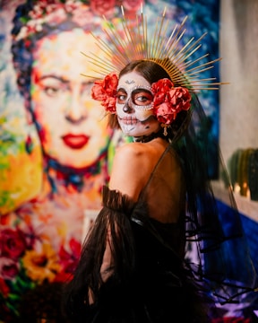 A woman is adorned in traditional Dia de los Muertos face paint and costume, featuring intricate skull designs and vibrant colors. She wears a black, sheer gown with bold red flowers around her head and a golden halo-like headpiece with spikes radiating outward. In the background, there is a vivid, colorful mural of a woman's face accented by floral motifs.