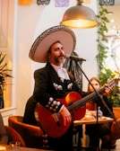 A person dressed in traditional mariachi attire is playing a guitar and singing into a microphone. The setting is warm and cozy with decorative banners hanging overhead and several plants in the background. A seated audience member listens attentively.