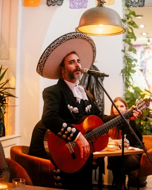A person dressed in traditional mariachi attire is playing a guitar and singing into a microphone. The setting is warm and cozy with decorative banners hanging overhead and several plants in the background. A seated audience member listens attentively.
