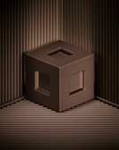A 3D-rendered brown cube with hollow square openings on its faces is positioned against a backdrop of ribbed, metallic brown panels. The cube appears to be floating above the base surface, casting subtle shadows that create depth. The overall appearance is minimalistic and geometric.
