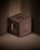 A 3D-rendered brown cube with hollow square openings on its faces is positioned against a backdrop of ribbed, metallic brown panels. The cube appears to be floating above the base surface, casting subtle shadows that create depth. The overall appearance is minimalistic and geometric.