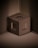A 3D-rendered brown cube with hollow square openings on its faces is positioned against a backdrop of ribbed, metallic brown panels. The cube appears to be floating above the base surface, casting subtle shadows that create depth. The overall appearance is minimalistic and geometric.