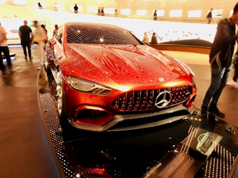 A sleek, red Mercedes concept car is displayed in a well-lit showroom with modern design elements. The car features a glossy finish with intricate detailing on the grille and headlights. Reflections and lighting give the car a futuristic appearance. People are walking around, observing the vehicle, which is prominently showcased on a platform.