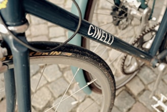 A close-up view of a bicycle focusing on the frame, wheel, and gears. The frame is dark blue with the word 'CINELLI' printed on it. The tire is slightly dusty, and the background is a cobblestone surface.
