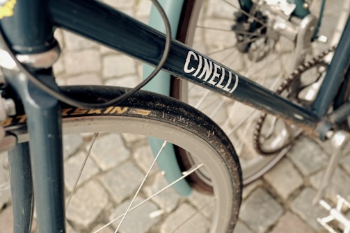 A close-up view of a bicycle focusing on the frame, wheel, and gears. The frame is dark blue with the word 'CINELLI' printed on it. The tire is slightly dusty, and the background is a cobblestone surface.
