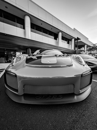 A high-performance sports car is parked in front of a modern office building with large windows and columns. The car has a sleek design with a prominent front grille and aerodynamic features.