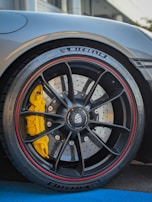 Close-up of a car wheel featuring a high-performance tire with MICHELIN branding and a detailed view of the brake disc and caliper. The caliper is bright yellow, providing a striking contrast to the black wheel with red trim. The wheel design includes multiple spokes and visible bolts, suggesting a sporty and premium design.