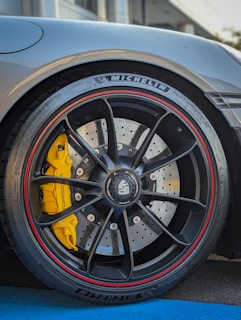 Close-up of a car wheel featuring a high-performance tire with MICHELIN branding and a detailed view of the brake disc and caliper. The caliper is bright yellow, providing a striking contrast to the black wheel with red trim. The wheel design includes multiple spokes and visible bolts, suggesting a sporty and premium design.