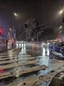 Street scene in rainy weather with reflections on wet pavement and lone figure holding umbrella.