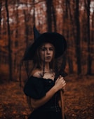 A person dressed in a witch costume stands in a forest during autumn. The background is filled with tall trees and fallen leaves, creating a mysterious atmosphere. The costume includes a black dress and a pointed hat, and the person holds a broomstick.