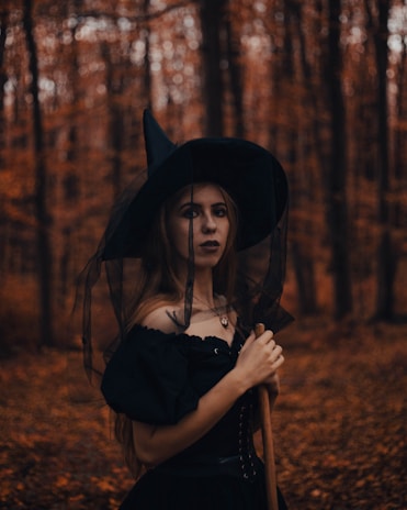 A person dressed in a witch costume stands in a forest during autumn. The background is filled with tall trees and fallen leaves, creating a mysterious atmosphere. The costume includes a black dress and a pointed hat, and the person holds a broomstick.