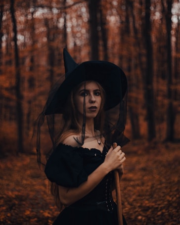 A person dressed in a witch costume stands in a forest during autumn. The background is filled with tall trees and fallen leaves, creating a mysterious atmosphere. The costume includes a black dress and a pointed hat, and the person holds a broomstick.